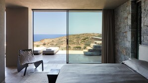 Quarto moderno com uma cama grande, área de estar com uma poltrona e vista para a paisagem costeira através de janelas do chão ao teto.