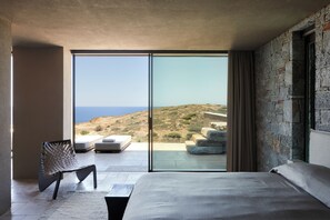 A modern bedroom with a large bed, a seating area with a chair, and a view of a coastal landscape through floor-to-ceiling windows. - The Tierras - Ryāki | One Bedroom Private Pool Villa (Mononaftis)