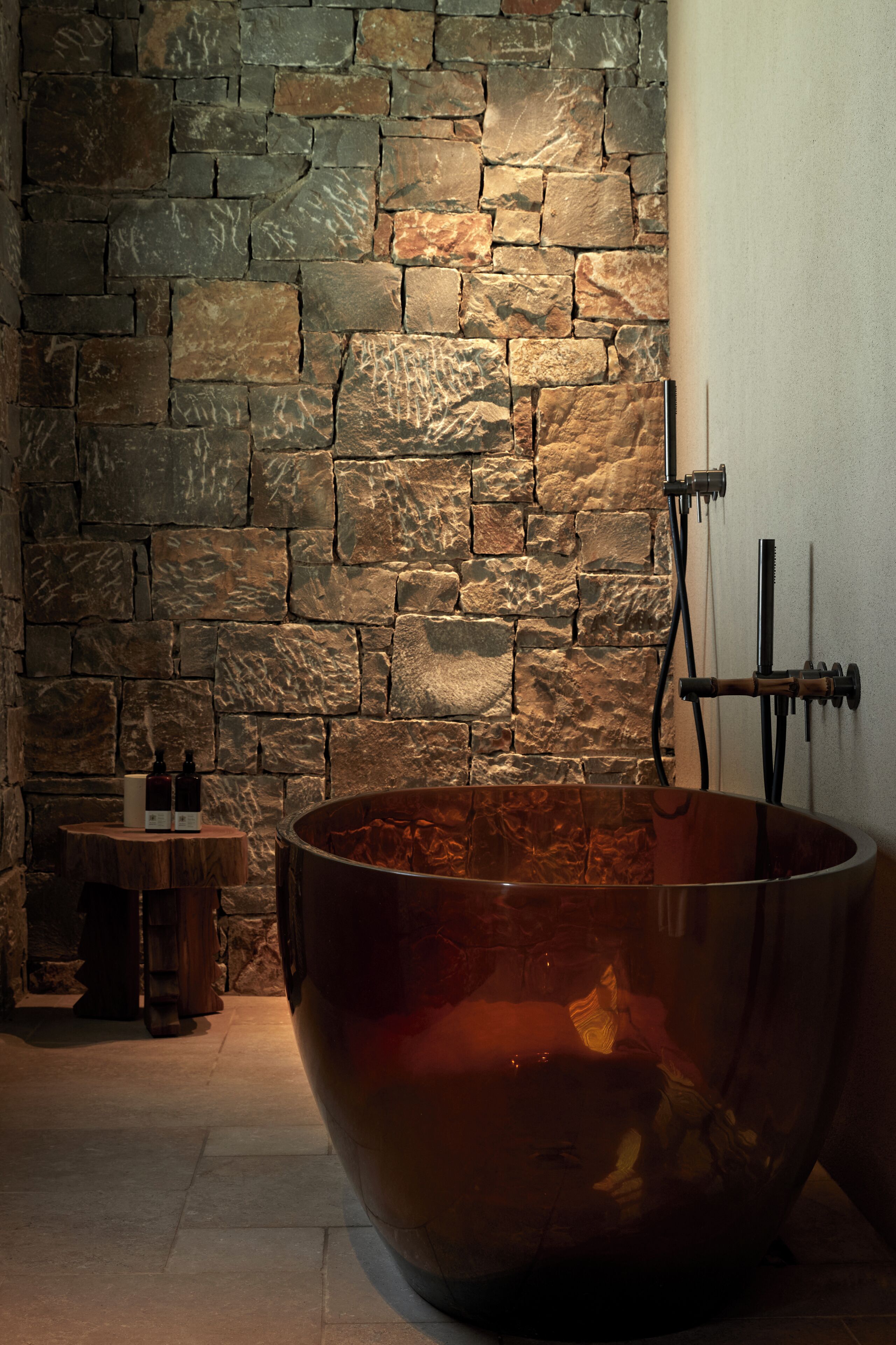 Un mur de pierres, une grande baignoire en cuivre et une petite table avec des bouteilles.