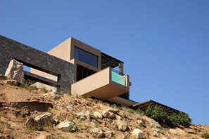 A modern building with a stone facade, a balcony with a glass railing, and a clear blue sky. - The Tierras - Ryāki | One Bedroom Private Pool Villa (Mononaftis)