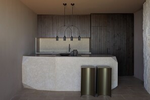 A modern kitchen with a curved island, pendant lighting, and a stone accent wall. - The Tierras - Ryāki | One Bedroom Private Pool Villa (Mononaftis)