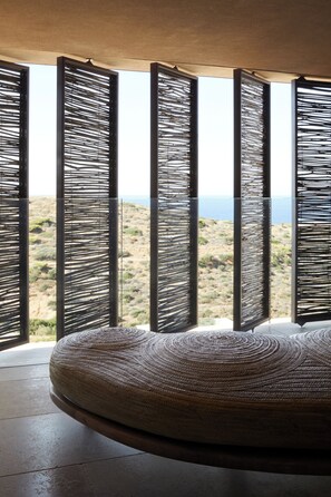 A room with a large woven ottoman and a view of a desert landscape through open louvered windows. - The Tierras - Phōs | 2 Bedroom Maisonette Private Pool Villa (Mononaftis)