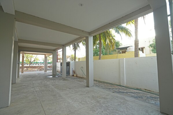 An indoor covered area with concrete flooring, white walls, and a view of palm trees and a yellow wall outside.