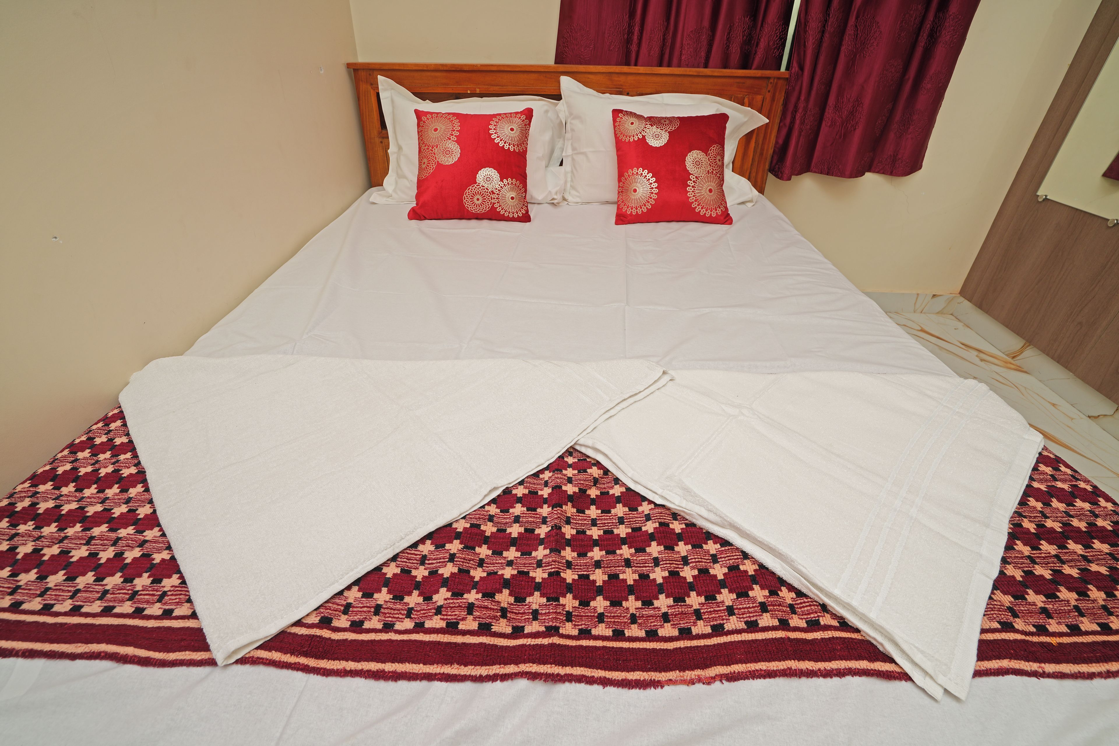 Una cama con ropa de cama blanca y dos almohadas rojas con motivos florales, un tapete rojo y beige con diseño, y un cabecero de madera.