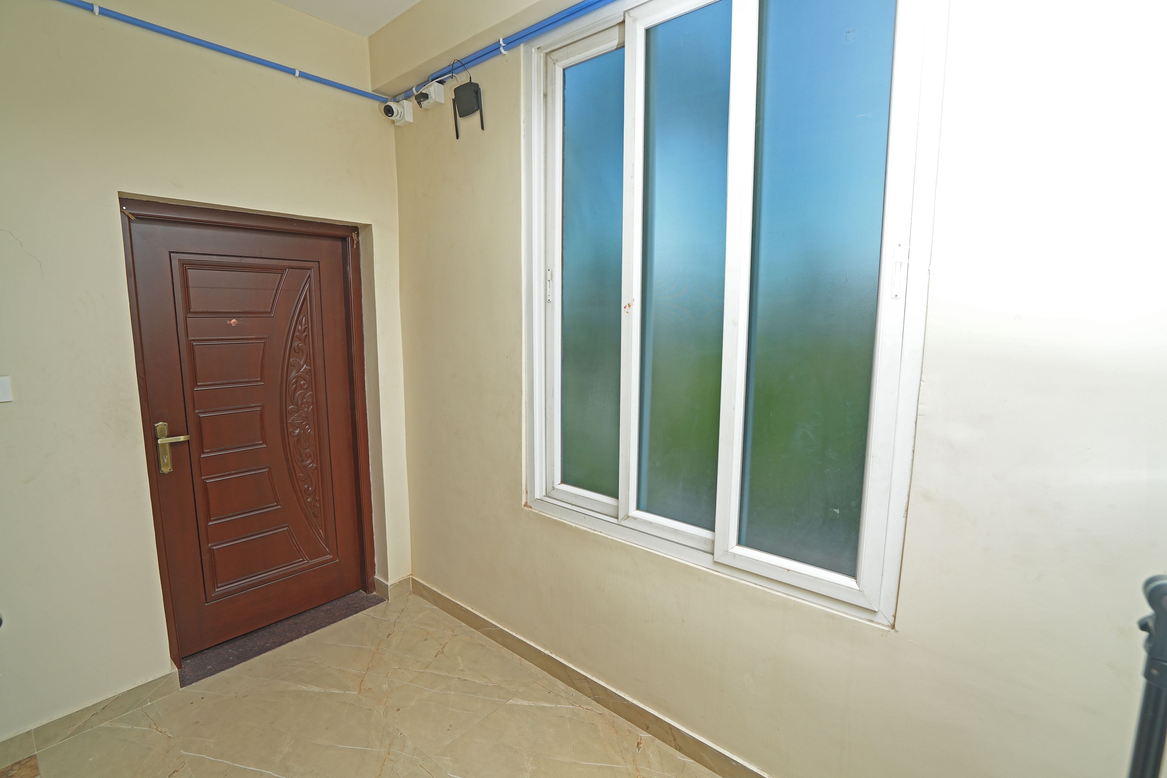 A hallway with a brown door, a window with a white frame, and a blue pipe running along the ceiling.