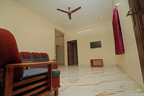 A living room with a sofa, a wooden coffee table, a ceiling fan, a TV mounted on the wall, and a door leading to another room.