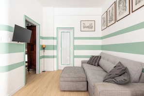 A room with a striped wall design, a grey sofa with cushions, a flat-screen TV mounted on the wall, and a door with a green frame. - Casa 3 Frecce a Corte Isolani by Wonderful Italy (Bologna)