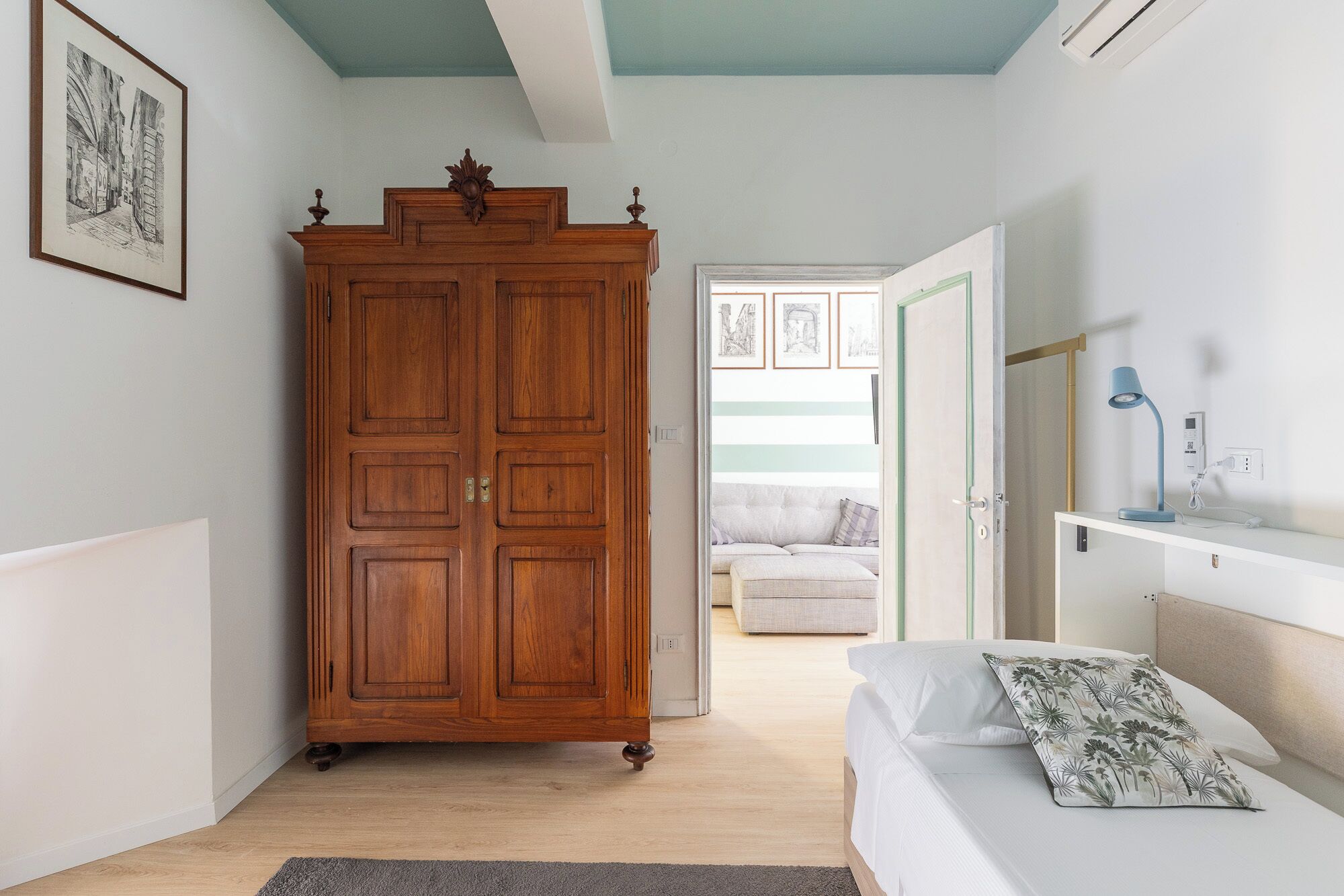 Een slaapkamer met een houten garderobe, een bed met een bloemenkussen, een nachtkastje met een lamp en een lijst met een afbeelding aan de muur.