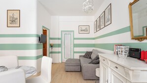 A room with a striped wall, a grey sofa, a white armchair, a wooden floor, and a white cabinet with books and a basket. - Casa 3 Frecce a Corte Isolani by Wonderful Italy (Bologna)