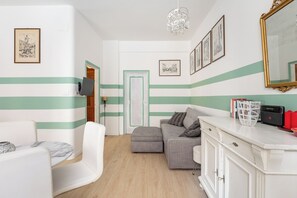 Ein Zimmer mit einer gestreiften Wand, einem grauen Sofa, einem weißen Sessel, einem Holzboden und einem weißen Schrank mit Büchern und einem Korb.