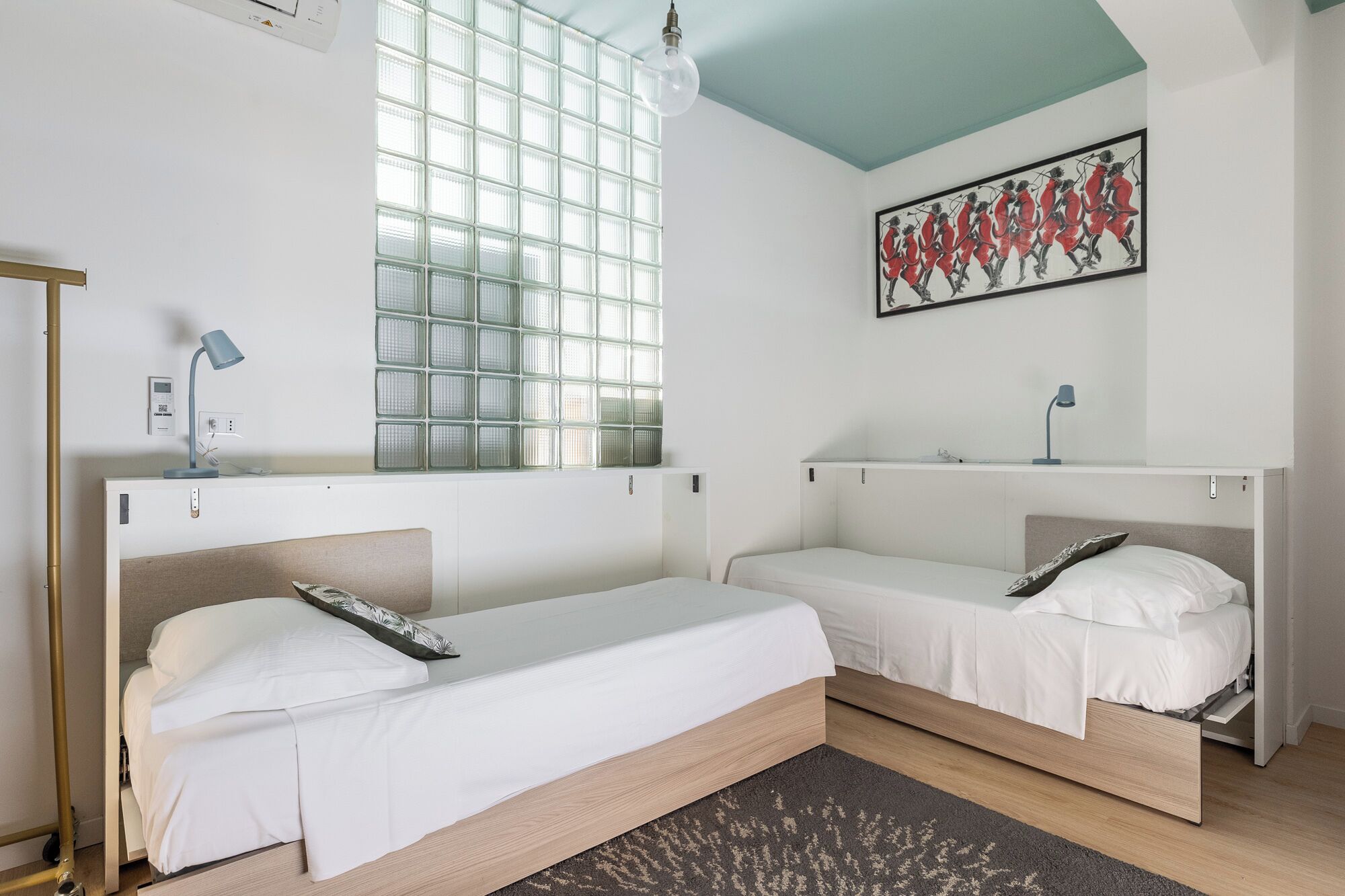 A room with two beds, a window with a view, a painting on the wall, and a desk with a lamp.
