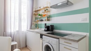A compact kitchen with a built-in washing machine, a coffee maker, and a shelf with books and decorative items. - Casa 3 Frecce a Corte Isolani by Wonderful Italy (Bologna)