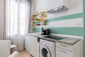 Eine kompakte Küche mit integriertem Waschtrockner, Kaffeemaschine und einem Regal mit Büchern und Dekoration.