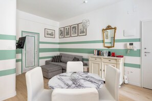 Una habitación con un sofá, una mesa de comedor con sillas, un aparador con libros y un espejo, y una puerta con cerradura.