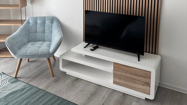Uma sala de estar moderna com uma televisão de tela plana sobre um console branco, uma poltrona azul claro e um painel de parede de ripas de madeira.