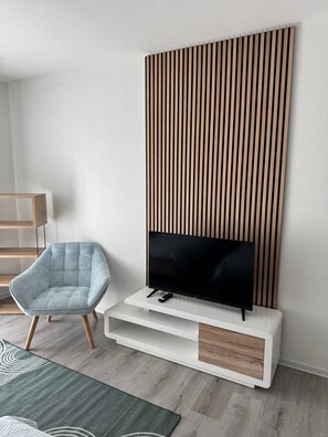 A modern living room with a flat-screen TV on a white console, a light blue armchair, and a wooden slat wall panel. - Apartment with Balcony, Kitchen & Wi-Fi (Balingen)