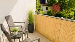 Uma varanda com móveis de vime, um vaso de planta e vista para uma casa e árvores.