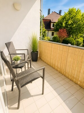 Ein Balkon mit Korbmöbeln, einem Blumentopf und Blick auf ein Haus und Bäume.