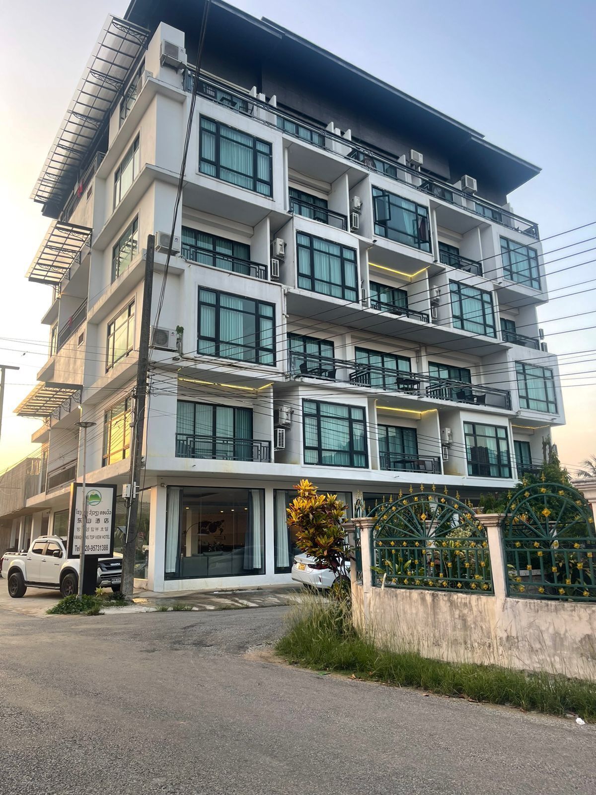 A multi-story hotel with a modern design, featuring balconies on each floor, large windows, and a gated entrance.