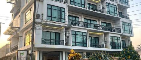 A multi-story hotel with a modern design, featuring balconies on each floor, large windows, and a gated entrance.