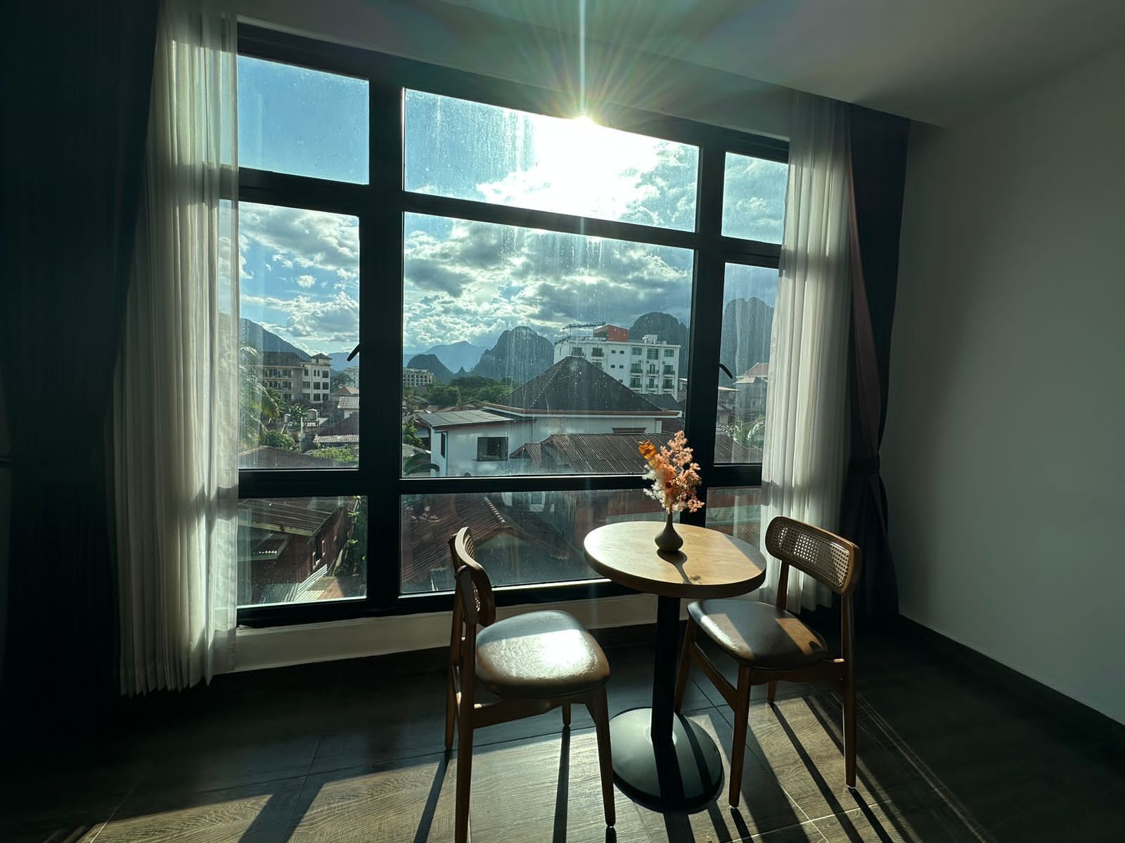 A room with a large window, a round table with two chairs, and a vase with flowers.