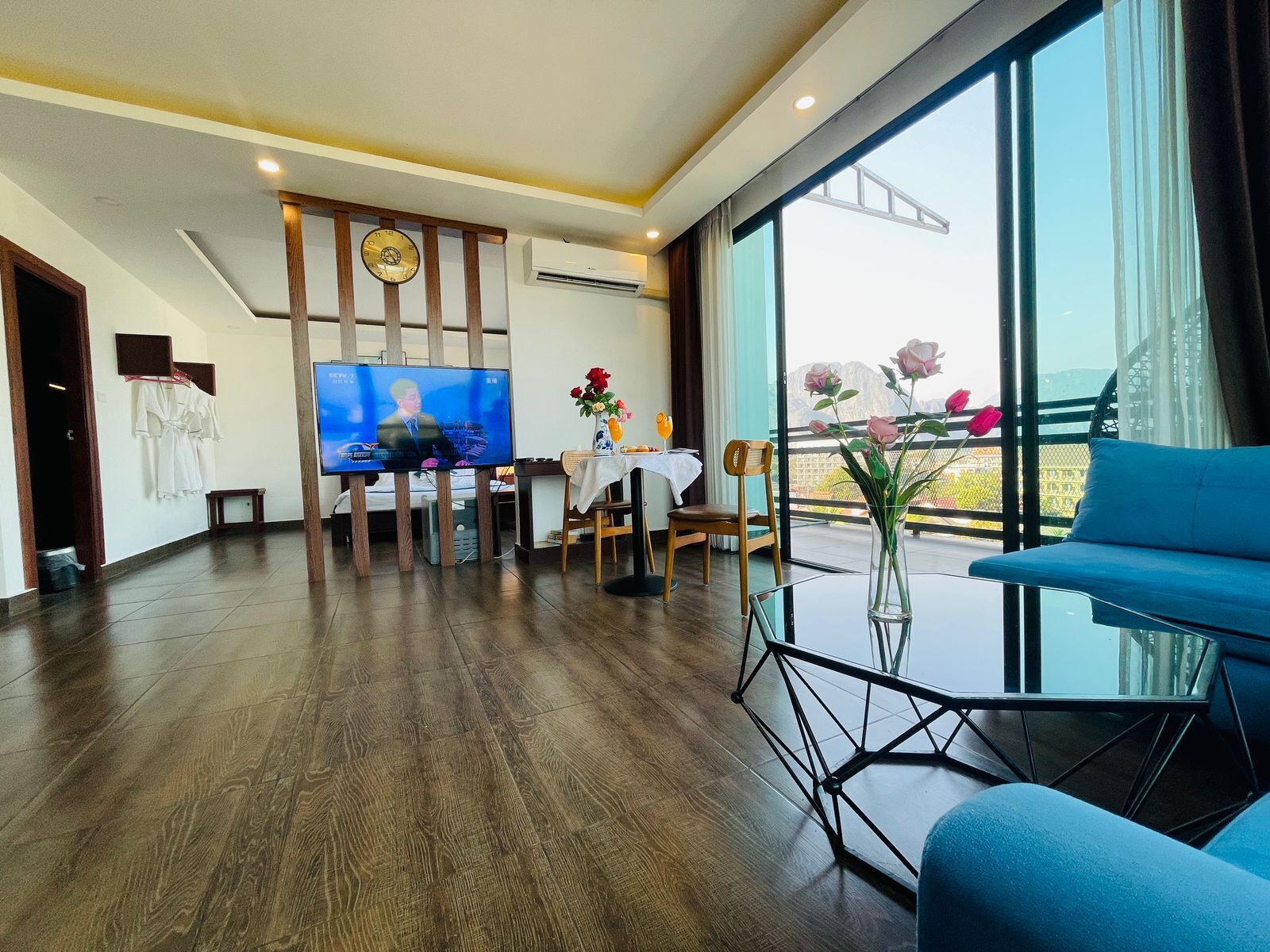 A modern hotel room with a large window, a flat-screen TV, a dining area with a table and chairs, and a blue sofa.