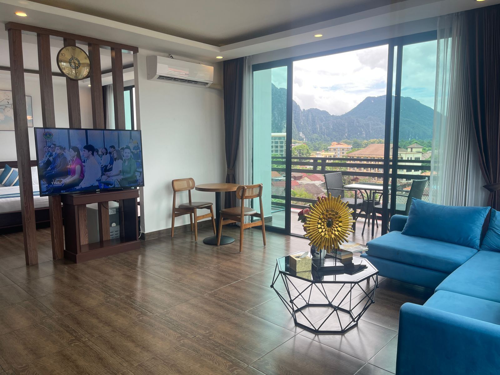 A modern hotel room with a large window, a television, a sofa, and a dining area.