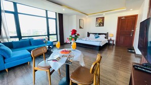 Una habitación de hotel con cama, sofá, mesa de comedor con sillas, televisor y un amplio ventanal con vistas a la ciudad.