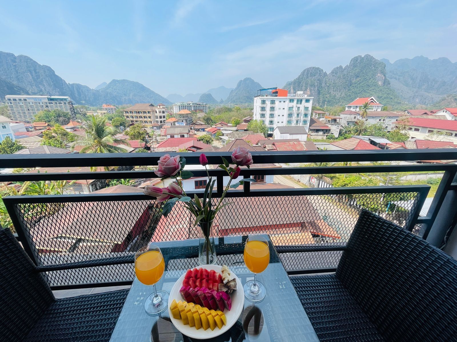 Un balcón con una mesa dispuesta para dos personas, que incluye un plato de fruta y dos vasos de jugo de naranja, con vista a un paisaje montañoso con edificios.
