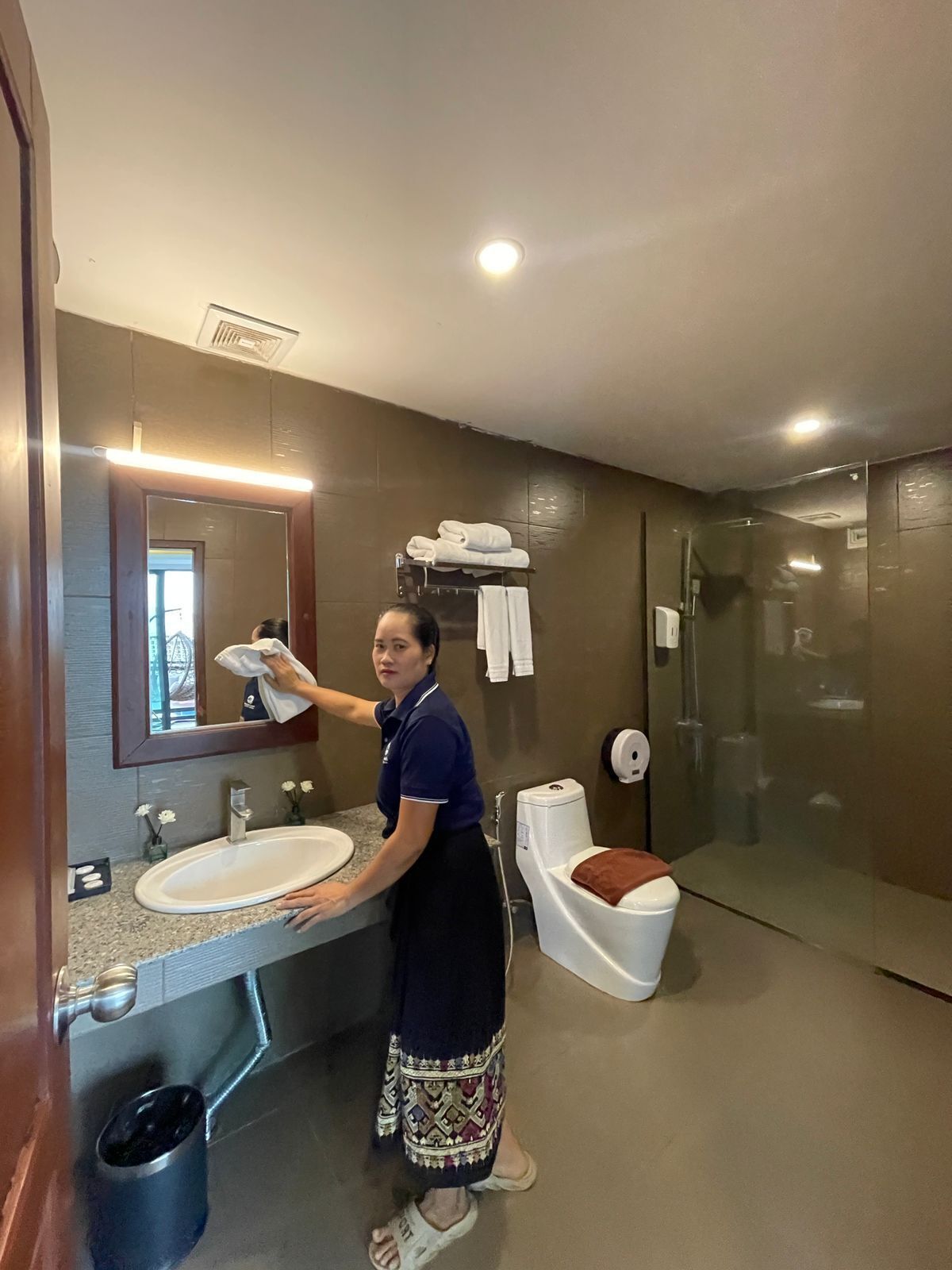 Une personne nettoie un évier dans une salle de bain moderne équipée d’un miroir, de toilettes et d’une douche à parois en verre.