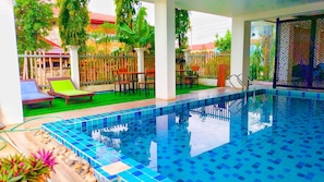 Une piscine aux carreaux bleus, entourée d’une terrasse en bois et d’un patio couvert aménagé avec des chaises et des tables.