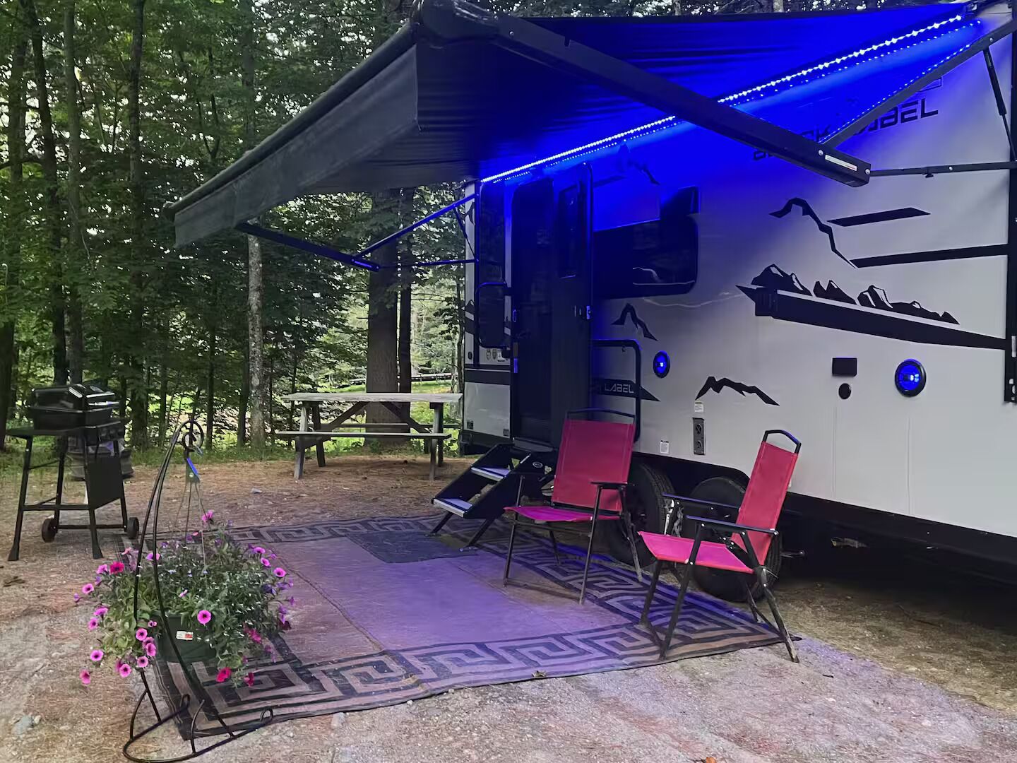 Un camping-car fermé, avec un éclairage intérieur bleu, une table de pique-nique et des chaises roses à l’extérieur.