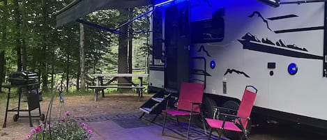 A covered RV with a blue interior light, a picnic table, and pink chairs outside.