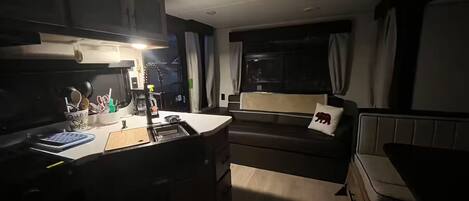 A compact kitchen with a microwave, sink, and various utensils. There is a small dining area with a sofa and a window with curtains.