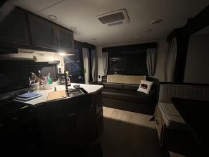 Ett kompakt kök med mikrovågsugn, diskho och diverse köksredskap. Det finns en liten matplats med en soffa och ett fönster med gardiner.