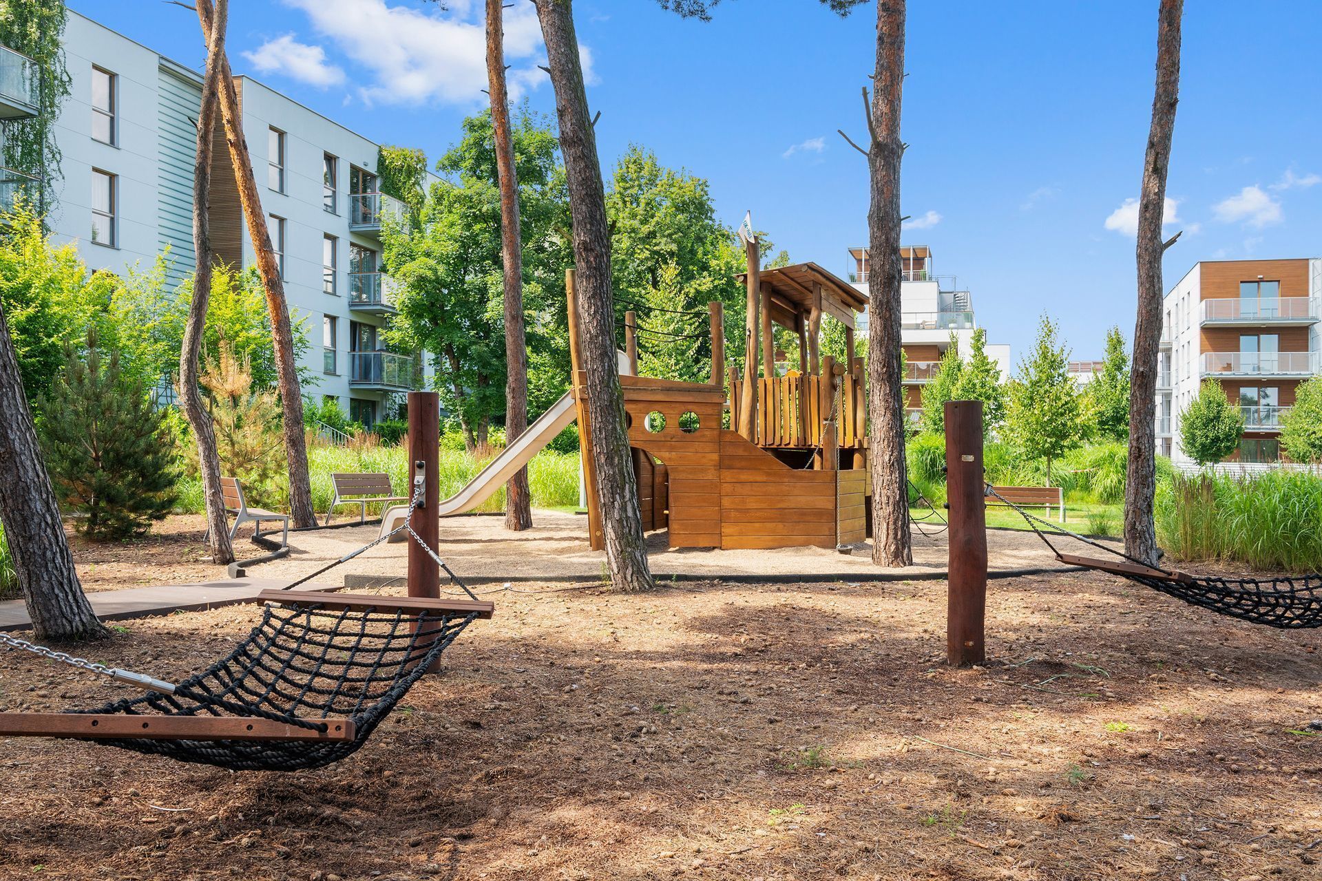 Un'area giochi con uno scivolo, altalene e una struttura in legno per arrampicarsi, circondata da alberi.