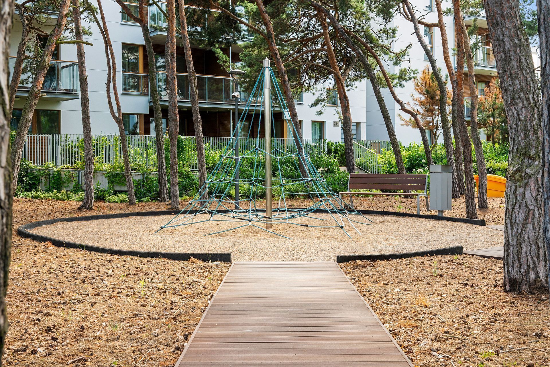 A wooden pathway leading to a playground with climbing structures, surrounded by trees and a building with balconies.