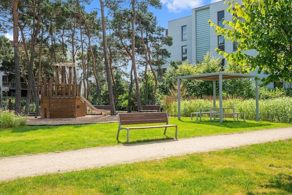 A playground with a slide and climbing structures, a bench, and a covered seating area in a park with trees and a building in the background.