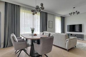 A modern living room with a dining area, grey upholstered chairs, a round wooden table, a sofa set, a flat-screen TV, and a chandelier.