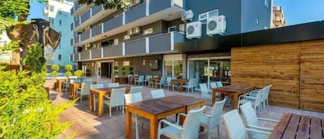An outdoor patio area with multiple tables and chairs, surrounded by a building with balconies and air conditioning units.