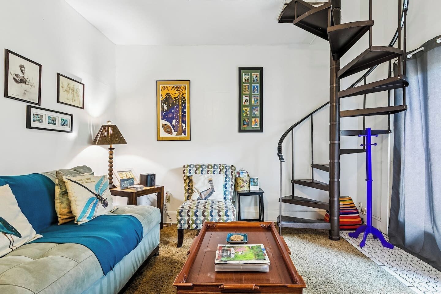 A living room with a sofa, a patterned armchair, a coffee table, a spiral staircase, and wall art.