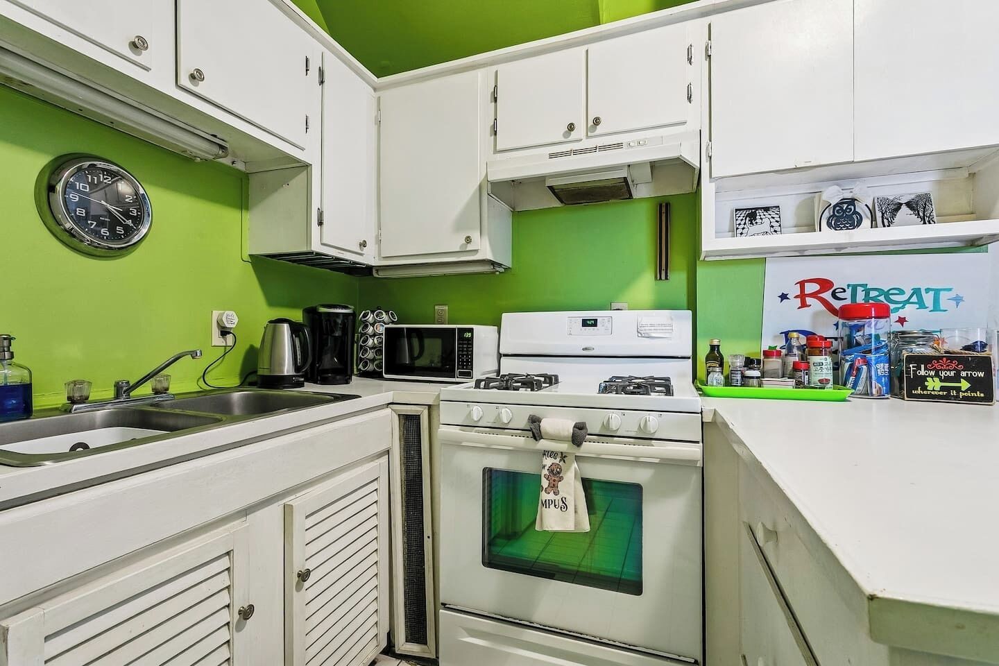 A kitchen with white cabinets, a microwave, a stove, and a sink. There is a clock on the wall and a sign that says 'Retreat'.