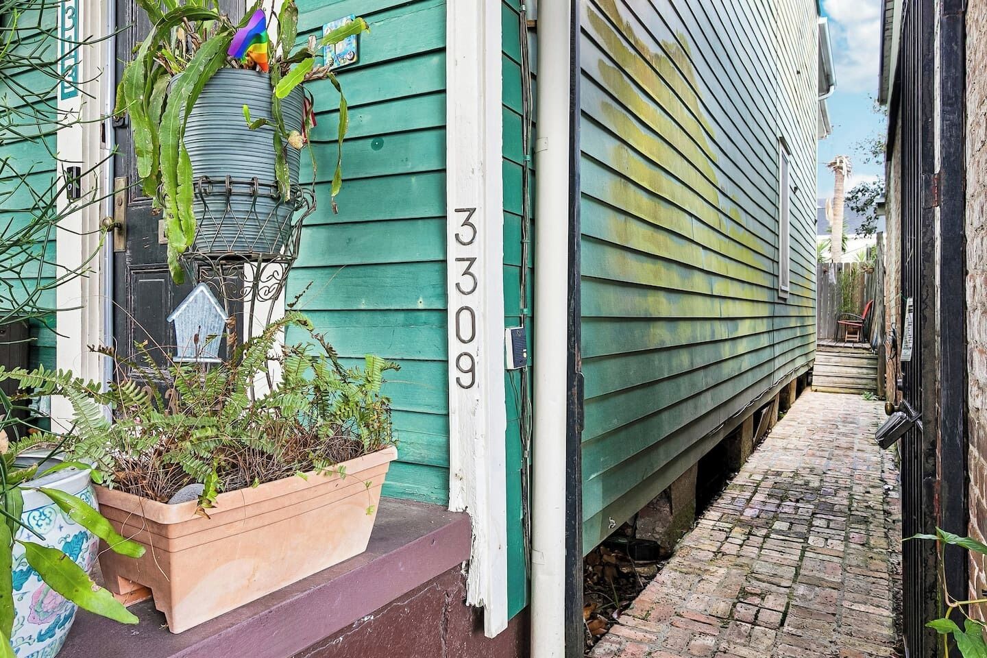 A narrow alleyway with a green house number 3309, a planter with plants, and a birdhouse on the porch.