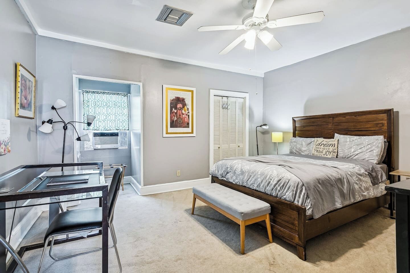 Un dormitorio con cama, escritorio, silla, banco, ventilador de techo y ventana con celosía decorativa.