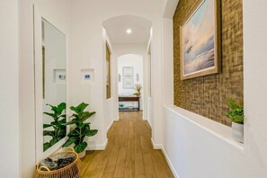 A hallway with a wooden floor, white walls, and a large mirror on the left. There are potted plants and a wicker basket on the floor. - All Inclusive- Nine Cities One Oasis/Game Room (Indio)