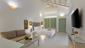 Una habitación de hotel moderna con sofá, una mesa de centro de vidrio con fruta, una cama con contraventanas verdes, un ventilador de techo y un televisor de pantalla plana.