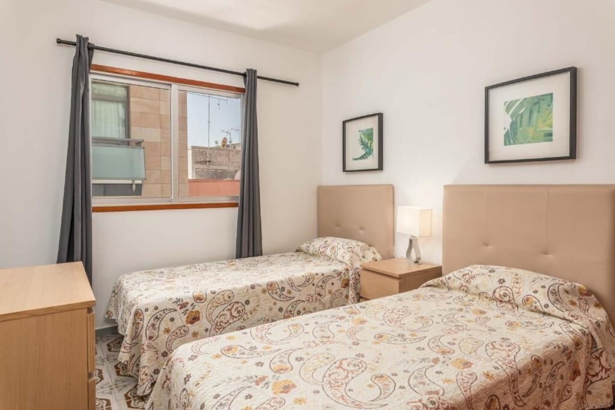 A hotel room with two beds, a window with curtains, a bedside table with a lamp, and two framed pictures on the wall.