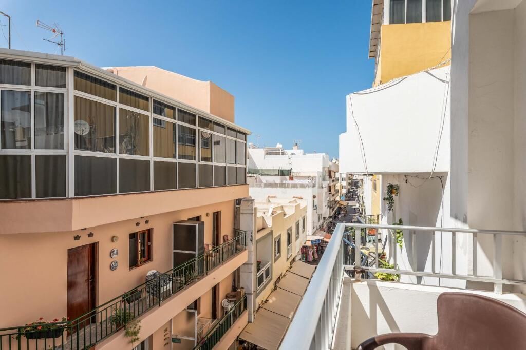 Un edificio residencial de varios pisos con balcones, cielo despejado y una calle estrecha.