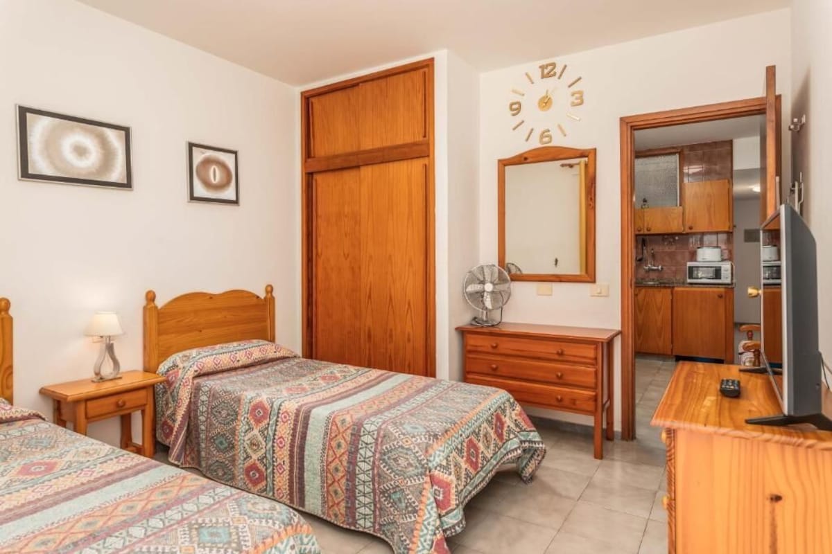 A bedroom with two beds, a wooden wardrobe, a desk with a mirror, and a kitchenette in the background.