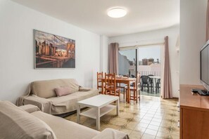 A living room with a sofa, a coffee table, and a dining area with chairs and a table. There is a television on a stand and a painting on the wall. - Edificio Los Playeros (Arona)